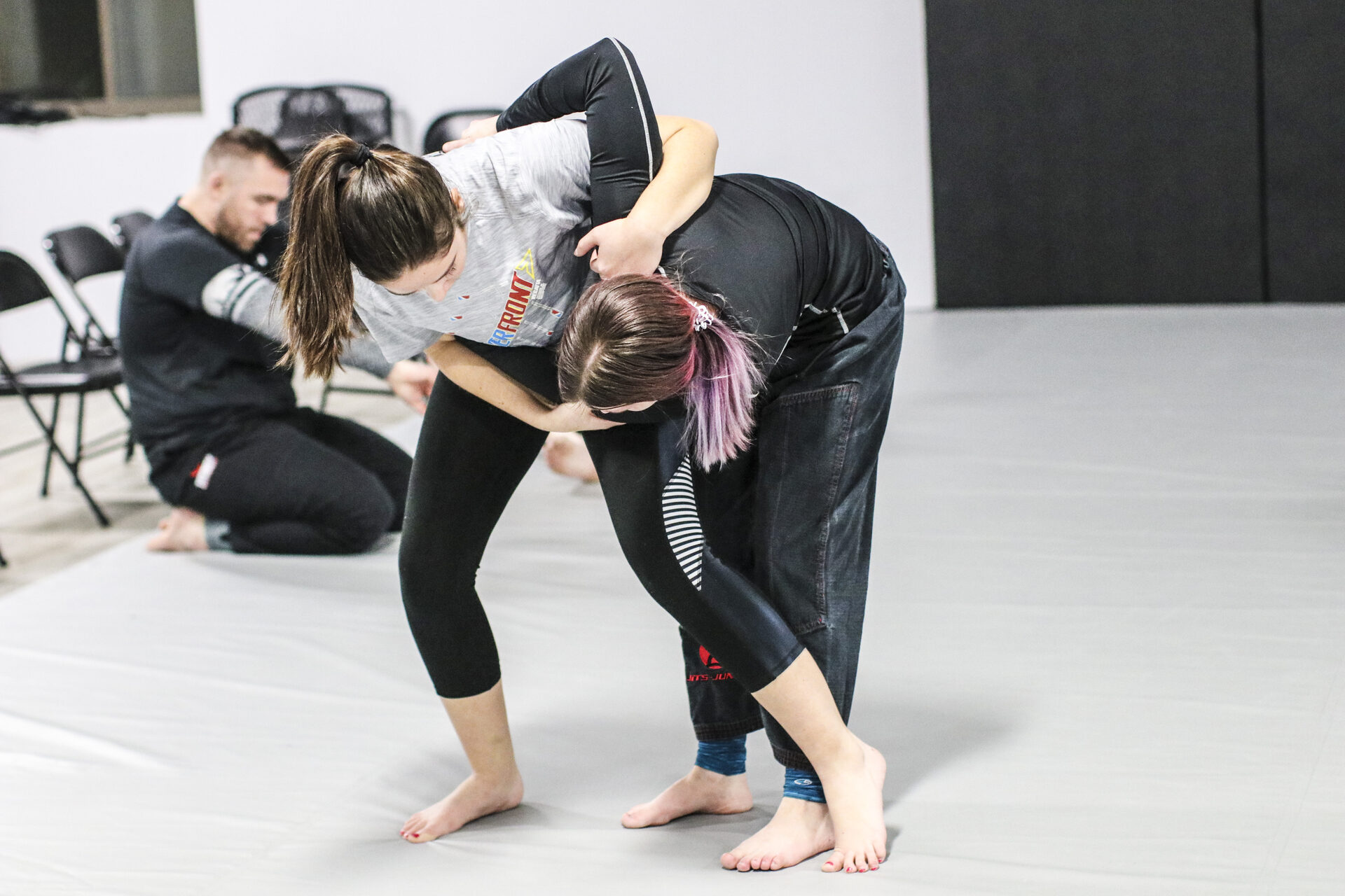 Two members in a standing tie-up, one bent forward stopping the other's entry, another member kneeling on the mat behind them