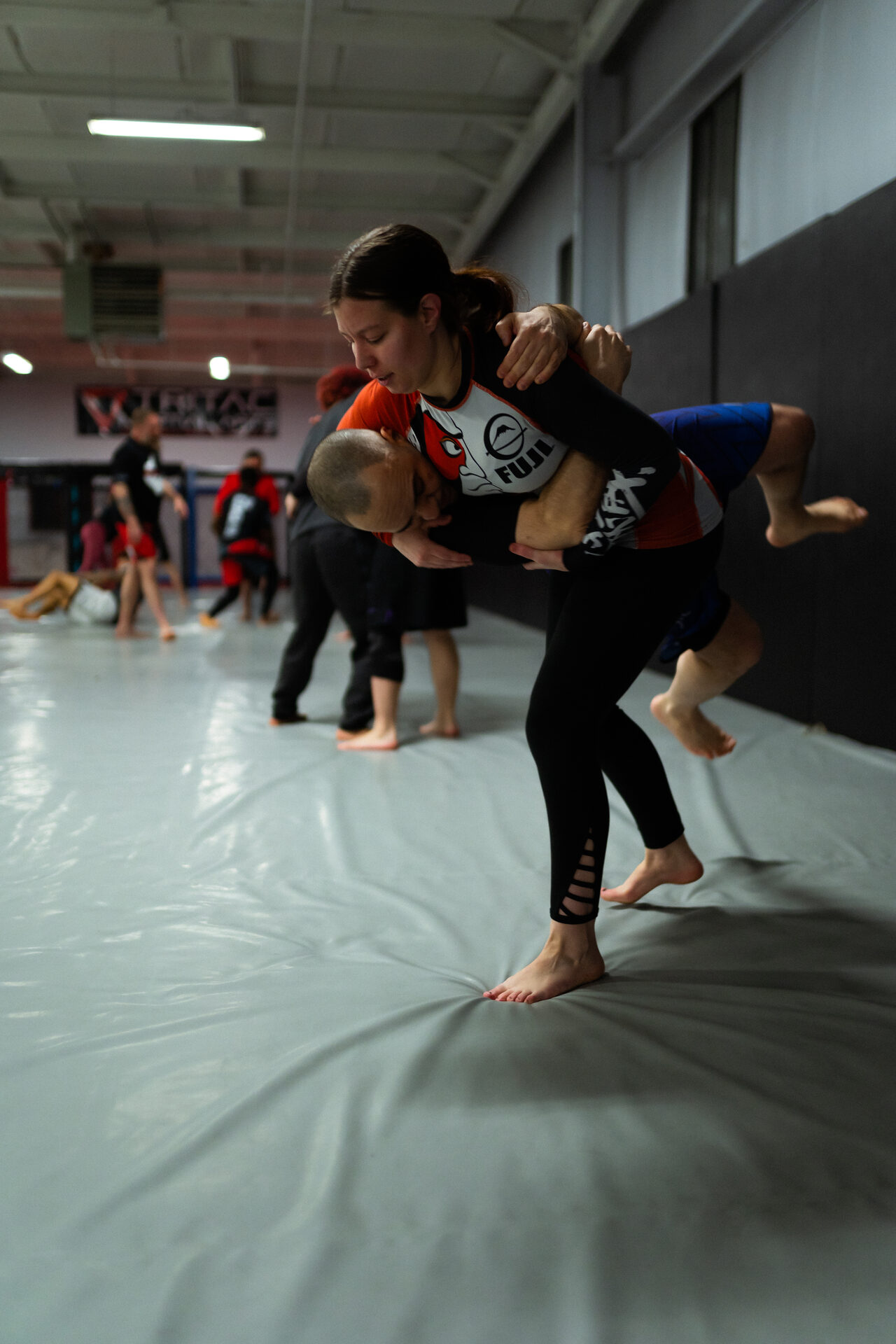 One member lifting her partner off the mat in a body lock during class drilling, other pairs working behind them