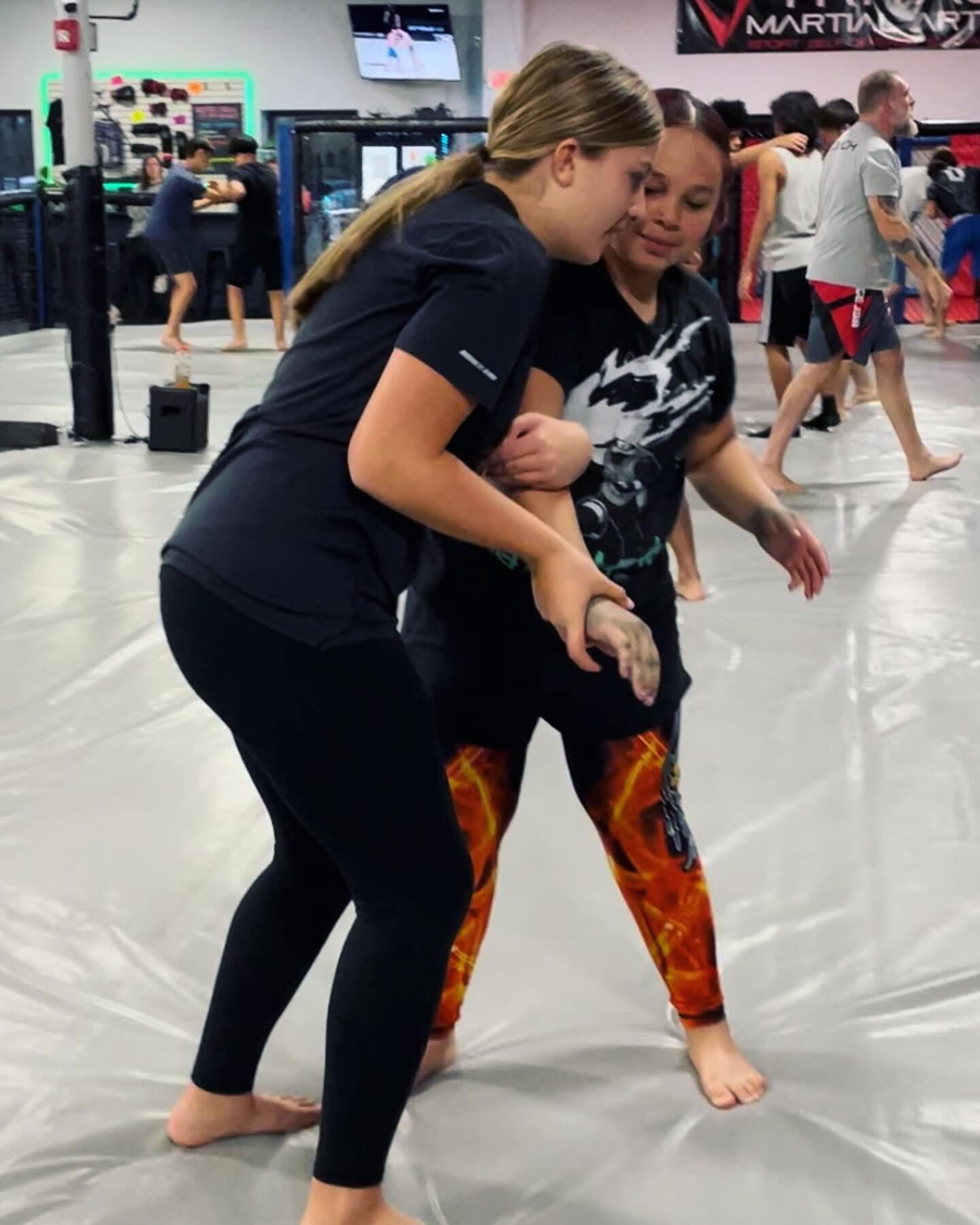 Two members clinched up on the mat, one working an underhook from the tie-up, a class training behind them
