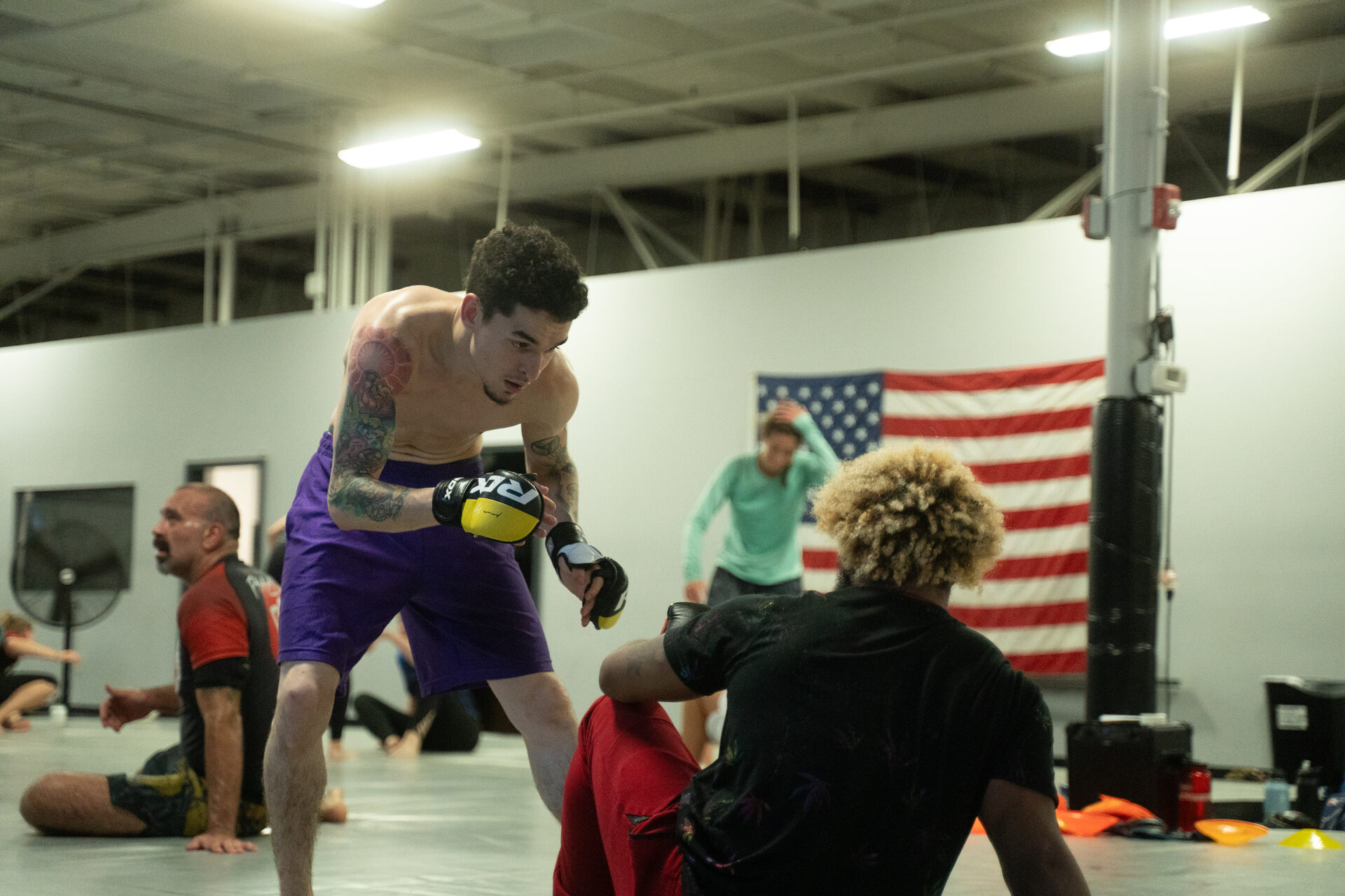 A live gloved round in progress on the mat, one member pressing forward while his opponent works to stop him and members watch from around the edge