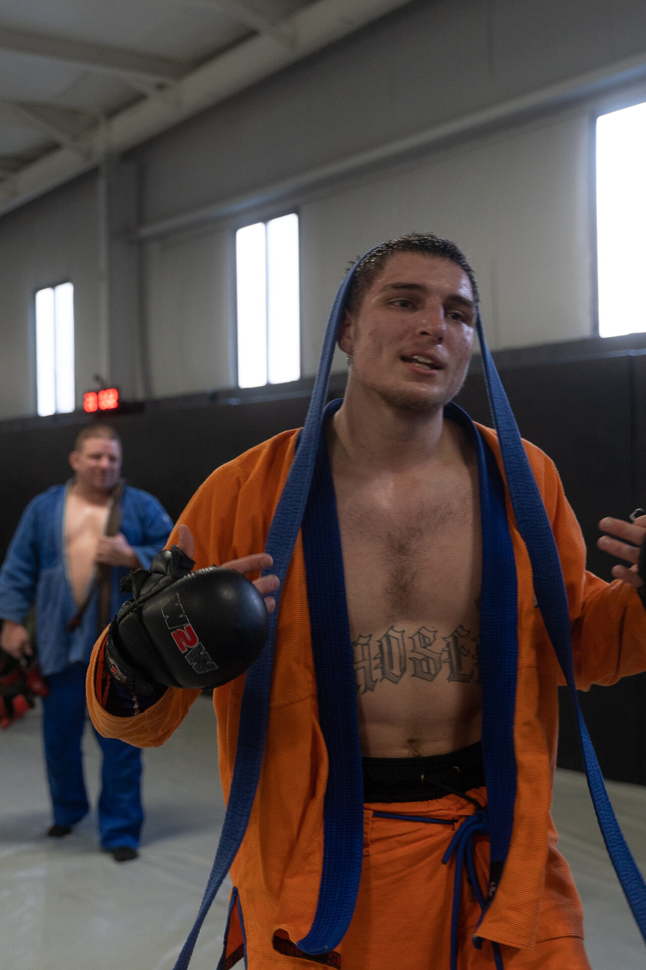 A fighter walking off after hard rounds, his gi open with his belt over his neck and sparring gloves still on